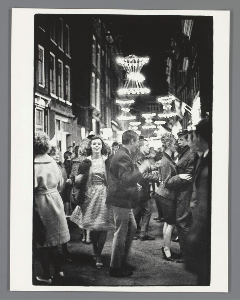 The image captures a lively scene from what appears to be an evening street festival in Amsterdam, specifically on the Nieuwmarkt during kermis. It is presented as a monochrome photograph that conveys a sense of movement and social interaction among people dressed in mid-20th-century attire. The focus centers around several couples dancing or walking through illuminated decorations suspended above them, which resemble oversized chandeliers with numerous light bulbs creating a radiant atmosphere.

The crowd includes both men and women wearing coats, dresses, and suits typical of the era's fashion trends for winter evenings. Some individuals are engaged in conversation while others dance, adding to the festive vibe captured by van der Elsken. The street is bustling with people moving in various directions, some blurred due to motion or focus effects, which contributes to a dynamic composition.

The photograph exudes an urban warmth and social engagement characteristic of kermis event