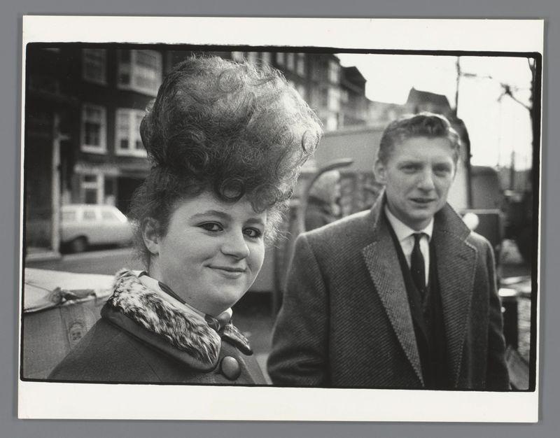 The image depicts a man and a woman standing side by side on what appears to be an urban street. The photograph is in black and white, suggesting it might be from the mid-20th century or earlier. The woman has voluminous hair styled upwards with curls at the ends, indicative of fashion trends popular around that time period. She wears a dark coat adorned with a fur collar which adds to her stylish appearance.

The man stands slightly behind her and is dressed in business attire consisting of a suit jacket, tie, and shirt; his expression appears more serious compared to hers. Both individuals are looking directly at the camera, suggesting they were likely unaware or unprepared for being photographed at that moment.

In the background, there's an urban setting with buildings lining what seems like a street where vehicles can be seen parked along it. The photograph has been scanned from physical prints and is presented in landscape orientation on a white border typical of Polaroid ins