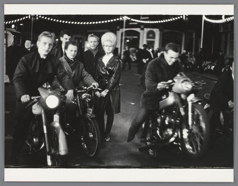 The image depicts a group of young people on bicycles and motorcycles, likely at an outdoor event or gathering. The scene is captured in black-and-white photography which adds to the nostalgic feel.

In the foreground, there are two individuals: one riding a motorcycle with a prominent headlight, turned towards another person who appears younger, possibly engaging in conversation. This individual has styled hair indicative of fashion trends from several decades ago and wears dark clothing.

Behind them is an older woman standing on her bicycle; she seems to be part of the group's focus as well. She too sports distinctive hairstyle that was popular during a specific period.

The background shows other people, some riding bicycles or motorcycles, suggesting movement through what appears to be decorated streets at night with lights adorning buildings and street poles. The overall atmosphere is lively yet casual.