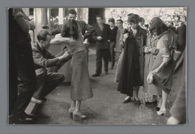 The image is a black and white photograph showing several individuals gathered outdoors, likely in an urban setting. A central figure appears to be assisting another person by adjusting their coat or dress around the waist area. The people are dressed in mid-20th-century attire typical of women's fashion with knee-length skirts and coats for men. Some onlookers stand nearby observing the interaction. In the background, a crowd can be seen walking through what seems to be an open-air market or public space filled with various activities. No explicit context is provided within the image itself regarding its specific location or event; however, it captures a candid moment of social exchange in a bustling environment.