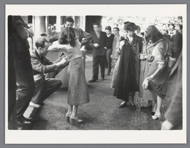 The image is a black and white photograph showing an outdoor scene with several people gathered. In the center, there's a woman facing away from the camera wearing a coat and holding what appears to be a piece of fabric or clothing against her body. She seems to be examining it closely.

A man on one side is kneeling down, writing something in a notebook while another person stands nearby taking notes with an open book. Several individuals stand around observing these actions. The people are dressed in coats typical of mid-20th-century fashion, suggesting the photo could date back several decades.

The setting appears to be casual and possibly related to some form of market or public event due to the number of bystanders and their engagement with each other's activities. There is a sense of everyday life captured at what might have been an exhibition or fair in Amsterdam during 1956, as indicated by the reference provided for this image.

The photograph has a candid quality, capt