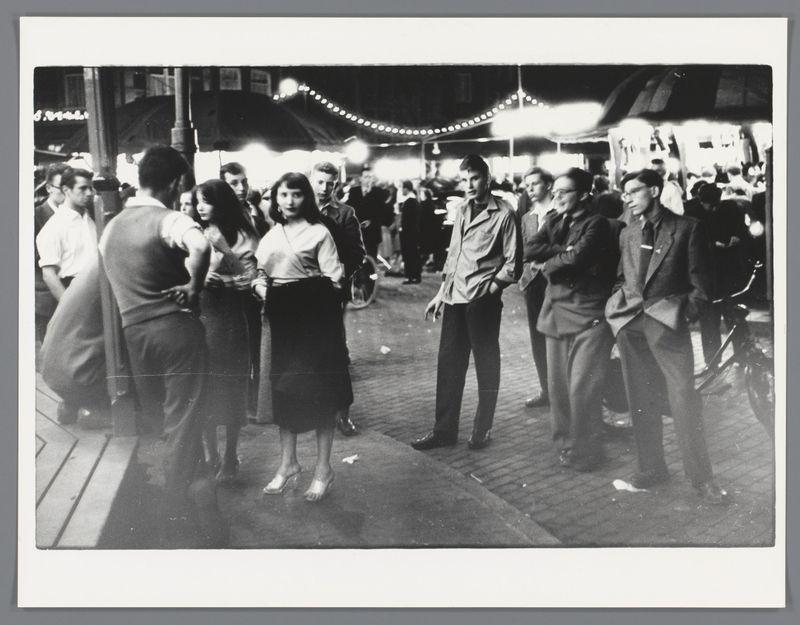 The image is a black and white photograph capturing a bustling night scene, likely from the mid-20th century. A group of people are gathered in what appears to be an outdoor market or fair with string lights illuminating the area. Various individuals are engaged in conversation; some stand close together while others maintain more distance between themselves. The attire suggests casual and semi-formal styles typical for that era, including dresses, suits, and jackets.

In the foreground, a woman stands out wearing a sleeveless blouse and skirt, interacting with someone off-camera to her right. Behind her are several men in conversation or looking around; one is notably smoking a cigarette. The background shows other attendees moving about, possibly engaging in activities like walking past stalls or watching performances. There's also evidence of bicycles parked near the edge of the scene.

The composition and lighting suggest it was taken candidly during an evening event with many 