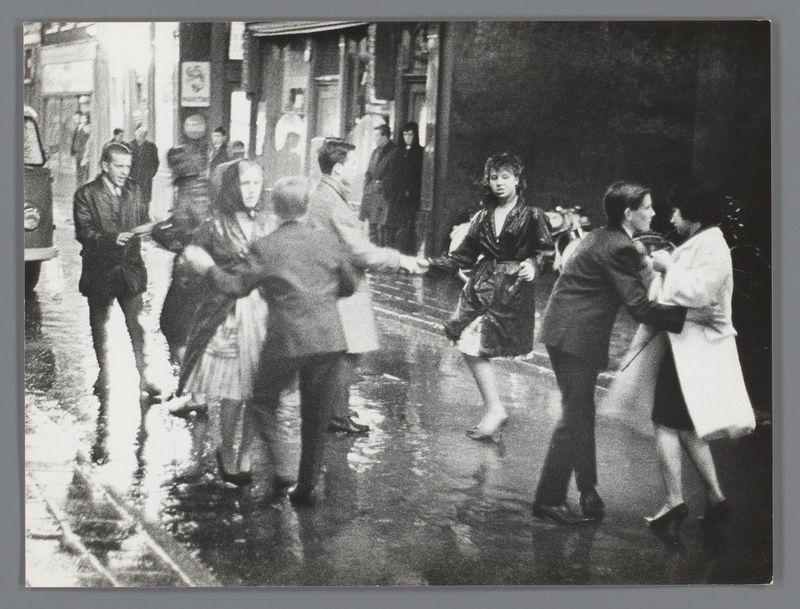 The image captures a lively street scene, likely in Amsterdam's Nieuwmarkt area during the festivities of a fair. It is rendered as a black and white photograph with high contrast, emphasizing textures like wet pavement reflecting light and puddles suggesting recent rain.

In the foreground, several individuals are engaged in dancing; their movements fluid and dynamic. A pair on the left side appears to be twirling around each other, arms linked and bodies swaying gently. The woman wears a long coat or dress with her hair pulled back from her face while the man is dressed more casually, suggesting an informal atmosphere.

To the right of this duo stands another couple in close embrace; they seem to pause momentarily before continuing their dance. Their attire indicates practicality for possibly wet weather conditions—jackets and coats suited for a brisk day or evening outdoors.

In contrast with these central figures are two other dancers on the far right, partially obscured by s