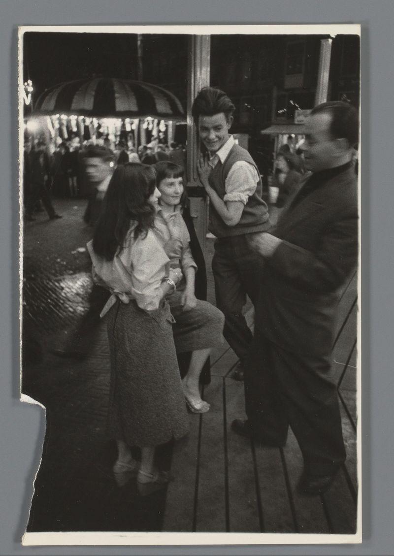 A black and white photograph depicting a lively scene at what appears to be an amusement park or fairground. In the foreground, four individuals are captured mid-conversation on a wooden walkway that suggests a carousel-like setting in the background.

The central figure is a young man with dark hair styled upwards, wearing glasses, a light-colored shirt under a brown vest, and patterned trousers. He seems to be animatedly engaging with his companions while holding what looks like a beverage cup or small container in one hand. To his left are two women: the first wears a sleeveless dress over a blouse paired with dark stockings; she gestures towards the central figure as if part of their conversation, displaying an expressive demeanor.

Beside her is another woman seated on a bench-like structure with ornate wooden detailing. She's clad in a light-colored top and patterned skirt, engaging more passively compared to the standing individuals beside her. The fourth person stands out p