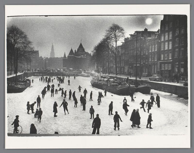 The image captures a historical winter scene of people engaging in outdoor activities on a snow-covered canal. Individuals are scattered across the frozen water, with some walking and others using what appear to be ice skates or perhaps early forms of rollerblades. The setting is urban, as evidenced by rows of buildings lining both sides of the canal. In the distance stands an iconic structure that resembles St. Nicholas Church in Amsterdam, suggesting this scene takes place there during a winter period when snowfall was not uncommon.

The photo is monochrome and shows signs of age with its grainy texture, which may indicate it's several decades old. The lighting seems to come from overcast skies or possibly street lamps, as the overall atmosphere has a muted quality typical of cold weather conditions at dusk or dawn during winter months in Europe. There are boats moored along the canal edges, and some individuals appear to be pushing sleds or skates.

The image is credited with "S