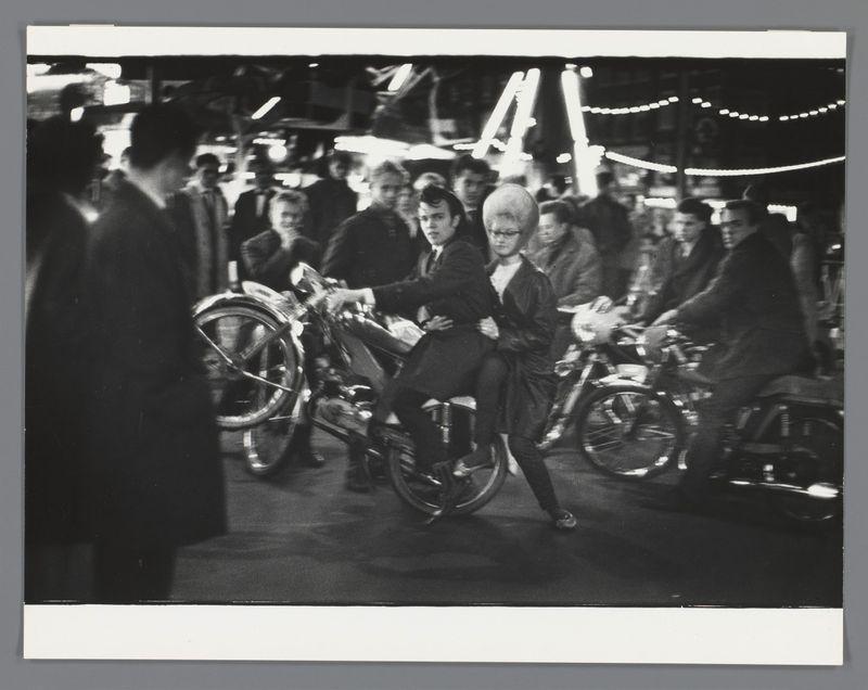 The image is a black and white photograph depicting several people riding motorcycles in what appears to be an outdoor evening setting. The focus of the photo centers on two women seated closely together on one motorcycle, with their posture suggesting they are about to start moving or perhaps have just come to a stop amidst traffic.

In addition to these riders, there is another person visible at the forefront wearing dark clothing and seemingly observing the scene, although not interacting directly. The background features other motorcycles and individuals who seem engaged in various activities typical of an urban environment during nighttime festivities such as lights from buildings or shops illuminating the area.

The composition captures a candid moment with a sense of immediacy, possibly hinting at the social dynamics between these subjects or their participation in some event or gathering. The overall atmosphere evokes a historical snapshot from Amsterdam's cultural life and