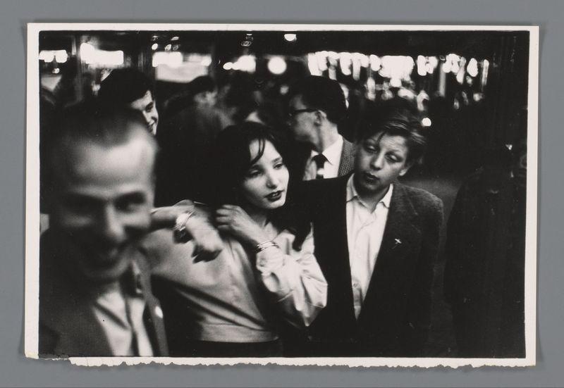 This black and white photograph captures a candid moment at what appears to be an evening social gathering or event. A group of people is seen in the background, with some individuals standing out due to their proximity to the camera lens and their expressive body language.

In the foreground, on the left side, there's a man who seems blurred as if he were moving quickly through the crowd. His expression is not clearly visible but suggests motion or surprise. In the center of the image stands another individual, possibly female based on clothing and hairstyle, looking directly at the camera with an expressive facial gesture that might be interpreted as speaking or reacting to something out-of-frame.

To her right, there's a person in dark attire wearing what appears to be a suit jacket with a pin on it. This figure has their head slightly tilted downward but is also facing towards the camera.

The background features additional figures who are somewhat indistinct due to the depth