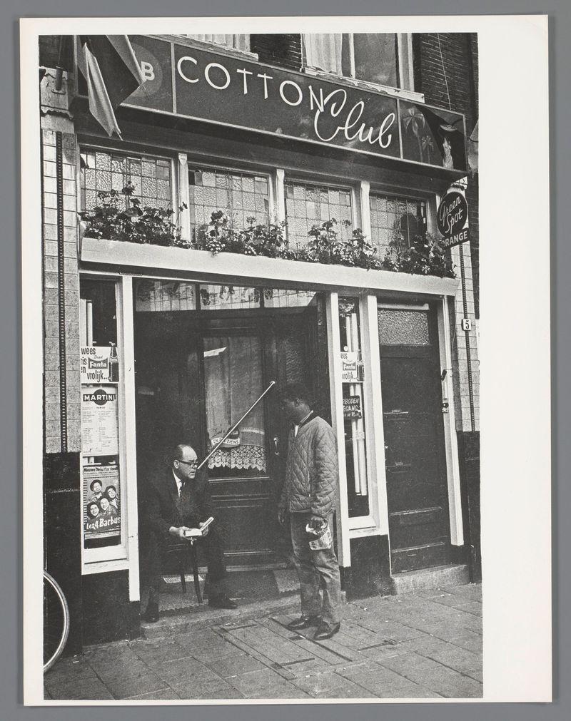 The image shows a black and white photograph of an urban street scene, likely from the early 1960s given the style. Two men are present; one is seated inside what appears to be a small establishment with large windows adorned with posters including 'MARTINI' and other items like bottles on display. The man sitting has his back towards us, facing an open door or entrance that leads into the building.

The second man stands outside in front of this establishment. He's dressed casually yet stylishly for what could be a cool climate; he wears glasses, a quilted jacket over a lighter top with pants and shoes suitable for urban walking. His posture suggests relaxed engagement as if waiting or pausing to interact momentarily before moving on.

The building is marked 'Cotton Club' in bold lettering above the entrance, suggesting it's either named such or was possibly used by that name at one point. Other signs indicate items like a "Green Spot" brand of beer and what seems to be an adverti