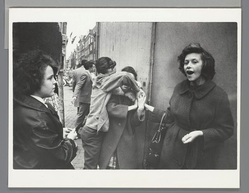 The image is a black and white photograph depicting an outdoor street scene. In the foreground, two women are captured in mid-conversation; one appears to be speaking with her mouth open while gesturing with her hand, wearing a dark coat. The other woman faces away from the camera, turned towards them, dressed in a light-colored jacket or overcoat and holding what looks like a small purse or bag.

In the middle ground, two more individuals are engaged in an interaction; one person is embracing another from behind, seemingly in surprise or alarm. This embrace occurs near the entrance of a building with visible signage. The scene captures a moment of everyday life on a bustling street, with various pedestrians and urban elements like buildings, walls, and possibly storefronts.

The photograph carries historical connotations as it seems to be taken during an era when such attire was common for women in public settings. No specific context or background story is provided within the ima