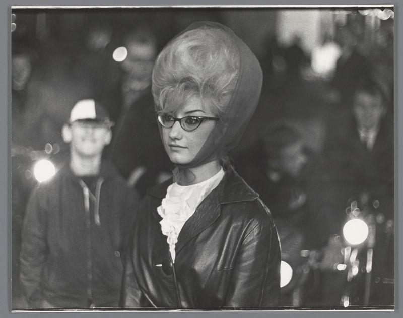 The image is a black and white photograph depicting an individual with voluminous, styled hair that reaches over their shoulders. They are wearing round-framed glasses and have on what appears to be a leather jacket layered over another garment with ruffled details at the neck area. The background suggests a busy environment with blurred lights and figures indicating it might be taken during nighttime or in low-light conditions. There's no explicit context provided, but the style of clothing hints at fashion from past decades. Additional information about this image can be found through its reference to Portret van Margriet Swart op de kermis op de Nieuwmarkt, Amsterdam by Ed van der Elsken dated 1956.