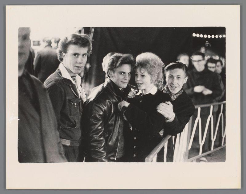 The image depicts a group of young people gathered closely together. There are five individuals visible, with four appearing to be boys and one girl among them. They stand against what looks like an outdoor fence or barrier railings at night or in low light conditions. The setting seems casual and possibly connected to some event as suggested by the presence of additional onlookers in the background.

The person second from the left is wearing a black leather jacket, while others have various styles including denim jackets and different colored shirts. Their hairstyles vary slightly but are indicative of a youthful and perhaps rebellious spirit typical of certain youth subcultures during that era. The boy with his arm around the girl's shoulder seems to be protective or friendly towards her.

The photograph is black-and-white, which lends it a timeless quality and suggests it may have been taken several decades ago when color photography was less common in casual snapshots. The foc